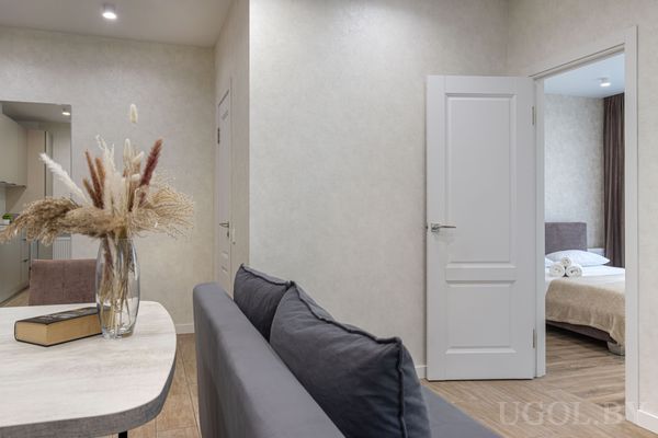 Интерьер с серым диваном, столом с вазой и книгой, на фоне — коридор, кухня и спальня с белым бельём и полотенцами.