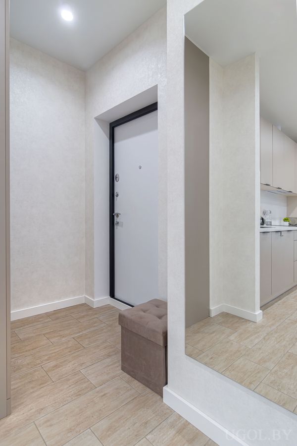 Минималистичная прихожая с белой дверью, коричневым пуфом и зеркалом, отражающим кухню с белой мебелью.