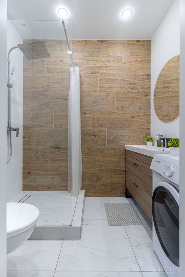 Современная ванная с душем, белой сантехникой, деревянной плиткой и встроенной стиральной машиной под раковиной.