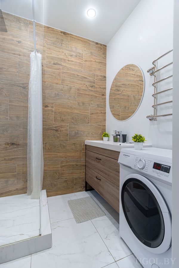 Современная ванная с душем за белой занавеской, деревянной плиткой на стене, круглым зеркалом над раковиной и стиральной машиной под полотенцесушителем.