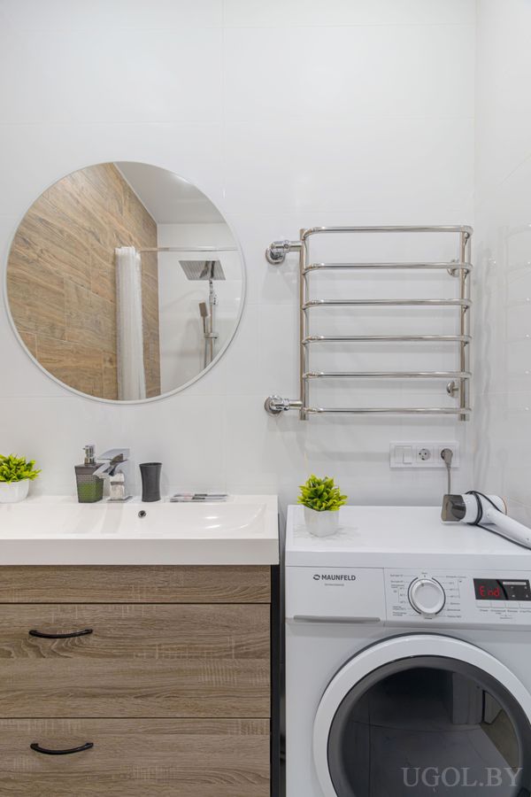 Чистая ванная с белой столешницей, деревянными ящиками, круглым зеркалом, стиральной машиной и настенным полотенцесушителем.