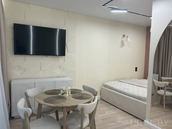 Интерьер студии с круглым столом на четыре места, телевизором на стене и кроватью с настенным светильником.