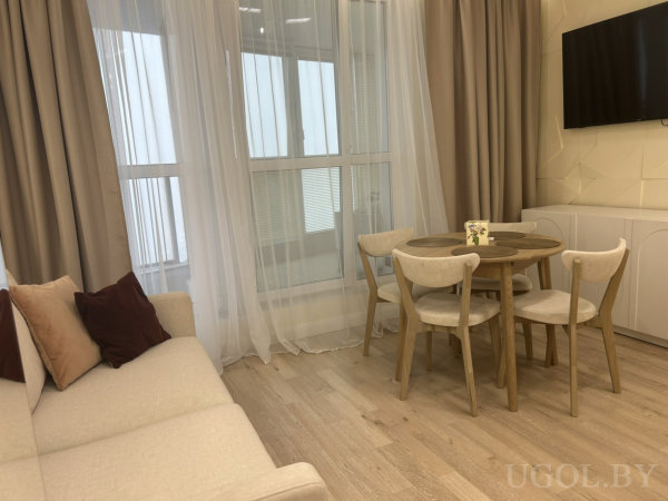 Светлая гостиная с бежевым диваном, круглым столом и телевизором на стене, рядом большое окно с шторами.