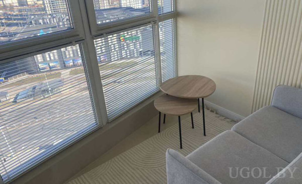 Серый диван у окна, два круглых столика, ковёр с волнами и панорамные окна с жалюзи в городской квартире.