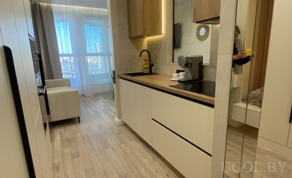 Компактная кухня с белыми фасадами, деревянной столешницей, чёрным краном и кофемашиной, рядом диван у окна.