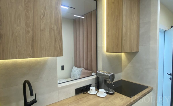 Минималистичная кухня с деревянной столешницей, кофемашиной, двумя чашками и зеркалом, отражающим мягкий уголок.