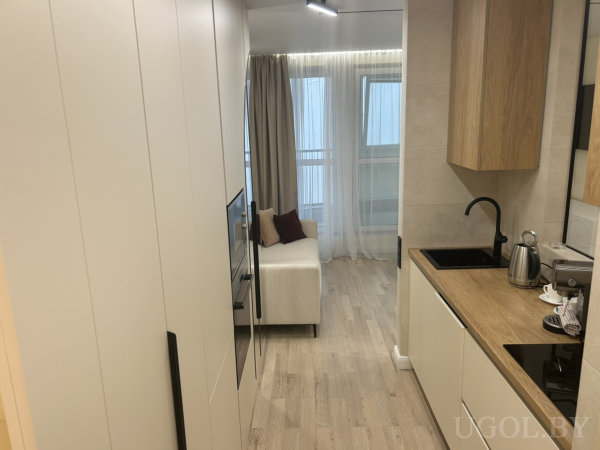 Белая кухня с чёрной сантехникой, встроенной техникой и деревянной столешницей, рядом диван и окно с тюлем.