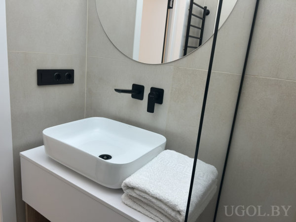 Современная раковина с чёрным настенным смесителем, стопкой полотенец и круглым зеркалом на бежевой плитке.