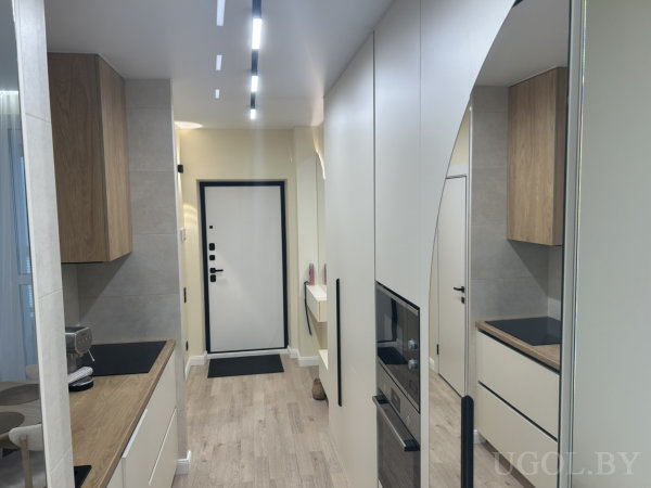 Узкая кухня с белыми фасадами, деревянными верхними шкафами, чёрной столешницей и зеркалом вдоль коридора.