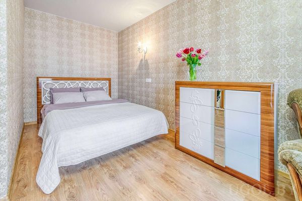 Современная спальня с бело‑серой кроватью, комодом с вазой тюльпанов и светлыми обоями с узором.