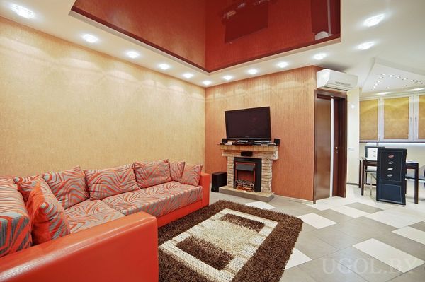 Красный угловой диван с узорными подушками, глянцевый красный потолок с подсветкой, телевизор над декоративным камином с каменной отделкой, плиточный пол и ковёр, вид на кухонную зону с барными стульями.