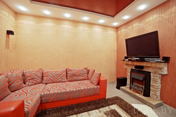 Красно-серый диван с узором, тёплое освещение в потолке, телевизор над камином с каменной отделкой и электрическим вставным очагом, ковёр с длинным ворсом, уютная современная атмосфера.