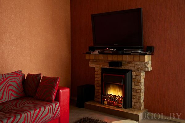 Красный диван с узорными подушками рядом с камином в каменной отделке, над ним телевизор и техника, стены в тёплых коричнево-красных тонах, создающих атмосферу уюта.