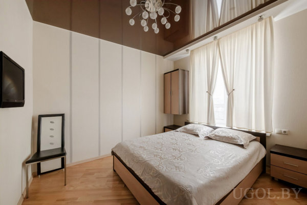 Двуспальная кровать с светлым покрывалом и тумбочками по бокам, шкаф у окна с тюлем, напротив телевизор и стул, потолок с глянцевой отделкой и люстра.