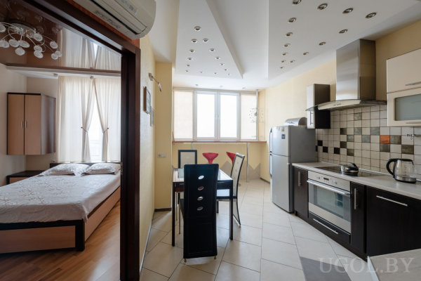 Кухня с современной техникой и плиткой в чёрно-бежевых тонах, обеденный стол со стеклянной столешницей и четырьмя стульями, рядом зона спальни с кроватью и шкафом, потолок с геометрической подсветкой.