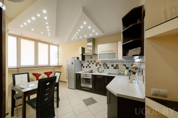 Тёмные нижние шкафы и белая столешница, плитка в серо-белой гамме, встроенная техника и вытяжка, обеденный стол со стеклянной поверхностью и чёрными стульями, у окна три красных барных стула.