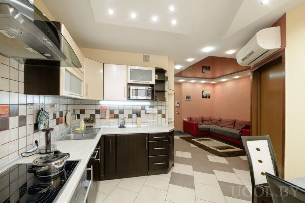 Современная кухня с тёмными нижними шкафами и светлыми верхними, белая столешница и плиточный фартук, встроенная техника и вытяжка. Рядом гостиная с красным диваном, ковром и тёплым освещением.