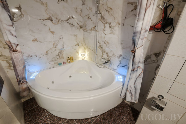 Угловая ванна с гидромассажем и встроенными светодиодами, мраморные стены, душевой шланг и шторы по бокам, на бортике туалетные принадлежности и светильник в форме сердца.