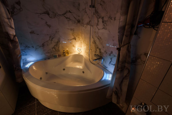 Белая угловая ванна с гидромассажем, голубая подсветка и свечи создают атмосферу уюта, мраморные стены, шторы по бокам и настенный светильник в форме сердца.