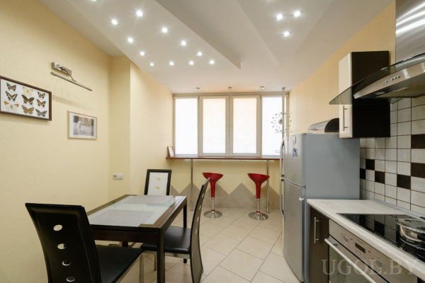 Просторная кухня с плиткой и светлыми стенами, стеклянный стол с чёрными стульями и картинами на стене, у окна барная стойка с красными стульями, современная техника и холодильник.