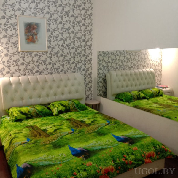 Яркое покрывало с павлинами и деревьями на фоне белого изголовья и черно-белых обоев, создаёт контраст с нейтральным интерьером. Над кроватью — абстрактная картина, рядом — зеркало и прикроватный столик.