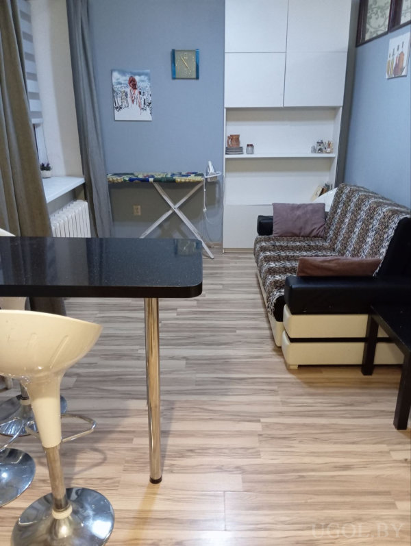 Пространство совмещает кухню и зону отдыха: барная стойка с белыми табуретами, окно с серыми шторами, гладильная доска у синей стены, рядом диван с узором и открытый стеллаж с декором.