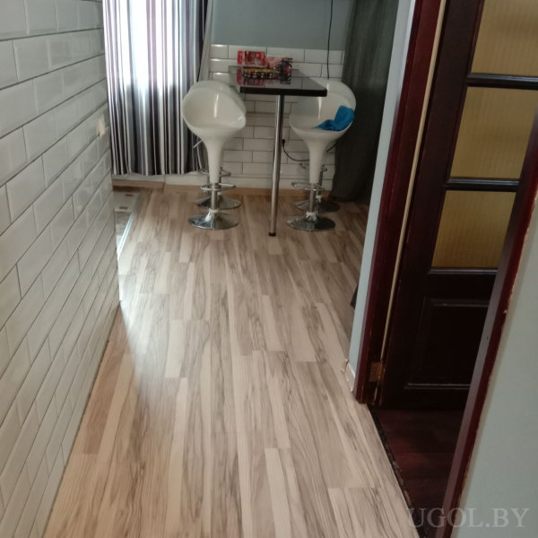 Небольшой стол с металлической ножкой и белыми барными стульями на фоне белой плитки и светлого ламината. На столе — красная коробка и мелочи. Окно оформлено полосатыми шторами, рядом — тёмная дверь.