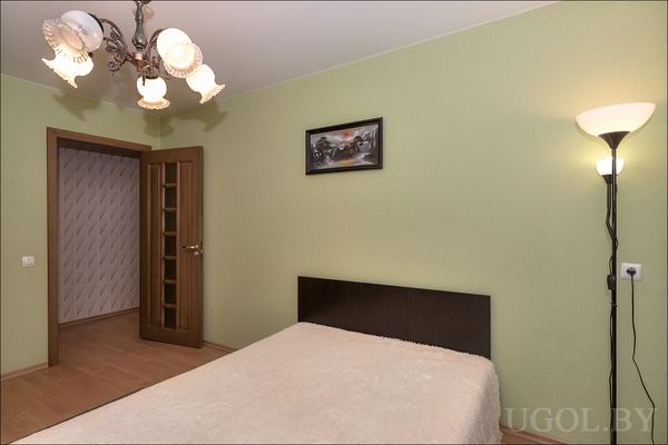 Комната с двуспальной кроватью, зелёными стенами и открытой дверью в зону с обоями. Чистый ламинат и потолочный светильник.