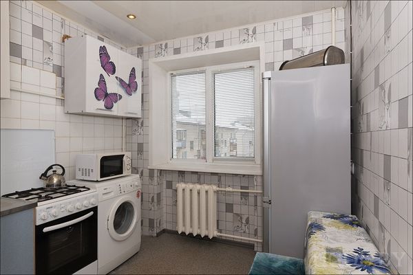 Компактная кухня с плитой, СМА и микроволновкой. Декор на шкафах, плитка с узорами и окно с жалюзи на город.