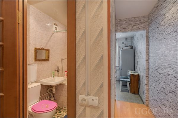 Вид на совмещённый санузел с розовой крышкой, зеркалом и переходом в кухню с обоями «под камень». Уютная планировка.