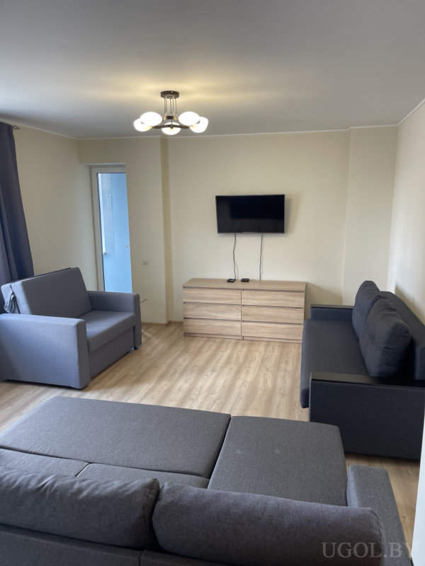 Комната с тремя серыми диванами, настенным телевизором над светлой тумбой, деревянным полом и окном с тёмными шторами.