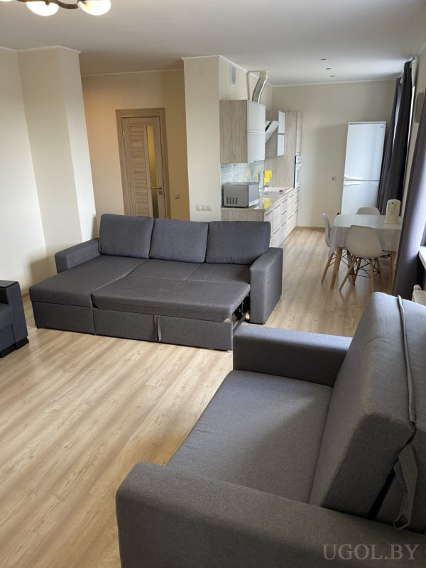 Комбинированная зона с двумя серыми диванами, обеденным столом с белыми стульями и кухней со светлыми фасадами и встроенной техникой.