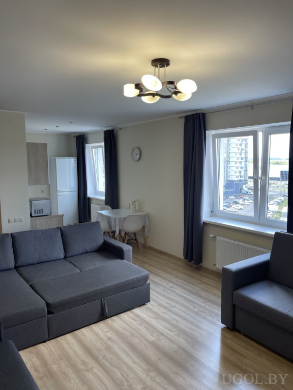 Комната с серым диваном и креслом, большим окном с тёмными шторами, обеденной зоной и кухней в светлых древесных оттенках.