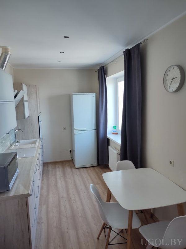 Светлая кухня с белыми шкафами, столом на двоих, тёмными шторами и холодильником у окна.