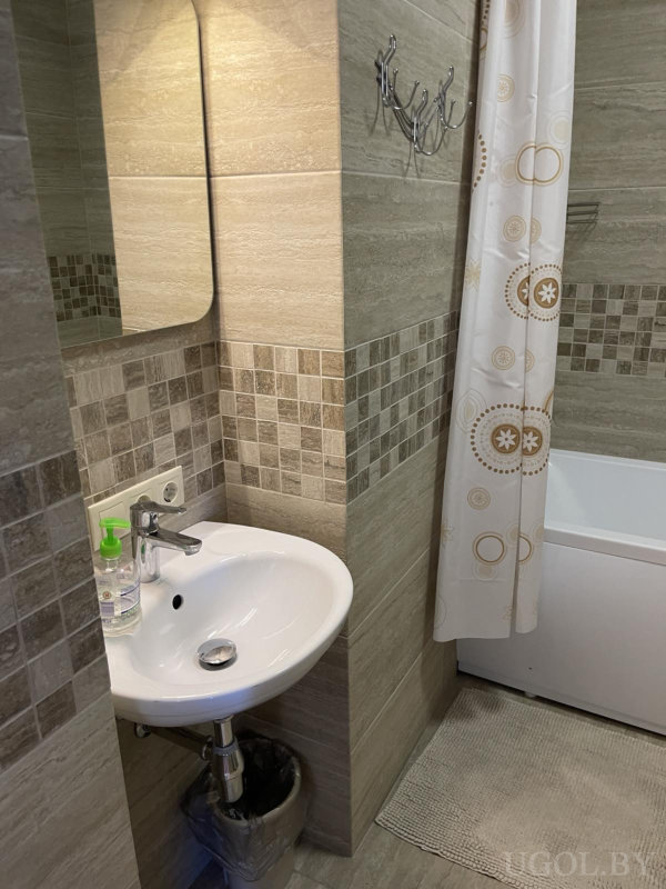 Современная ванная с раковиной, зеркалом, ванной с узорной занавеской и бежевой плиткой.