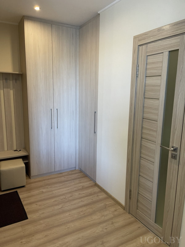 Светлая прихожая с угловым шкафом, панелью с крючками, небольшим пуфом и дверью в едином деревянном стиле.