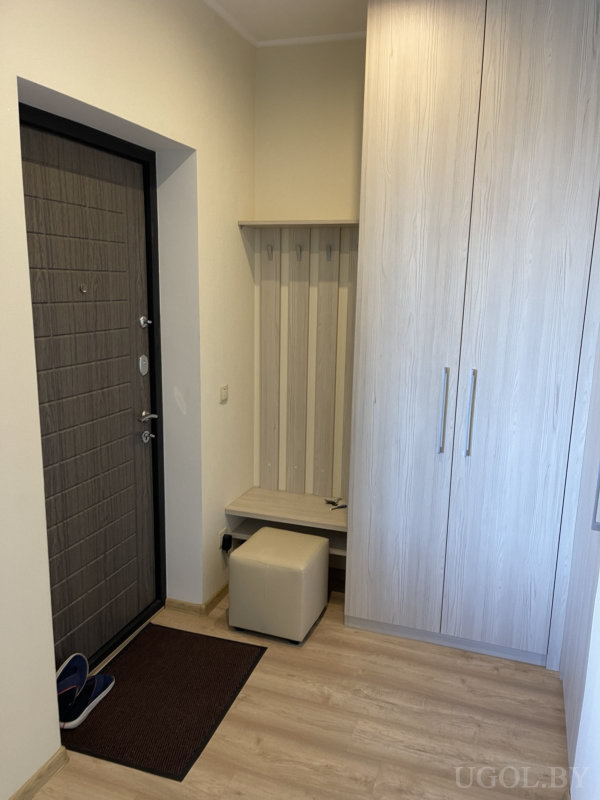 Компактная прихожая с тёмной входной дверью, ковриком и тапочками, настенной панелью с крючками, мягким пуфом и высоким шкафом из светлого дерева.