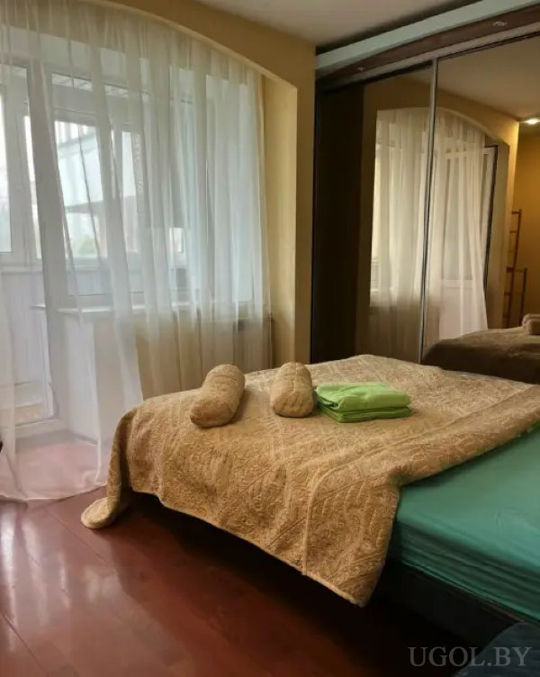 Небольшая спальня с двуспальной кроватью, покрытой бежевым покрывалом, на котором лежат свернутые полотенца. У окна — прозрачные белые шторы, справа — шкаф с зеркальными дверцами.