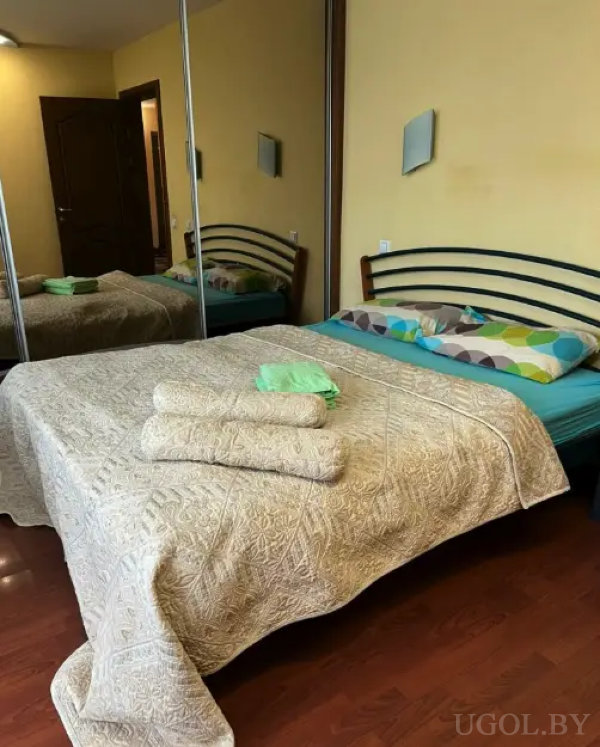 Кровать с бежевым покрывалом и цветными подушками, металлическое изголовье, зеркальный шкаф и теплый деревянный пол создают атмосферу уюта.