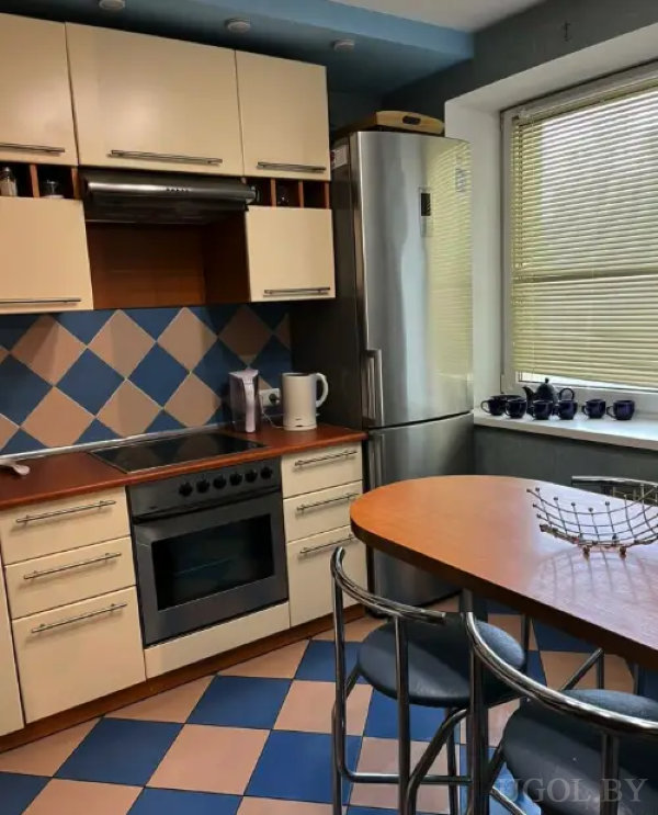 Интерьер с плиткой в сине-бежевом ромбовидном узоре, круглым деревянным столом, металлическими стульями и коллекцией кружек на подоконнике.