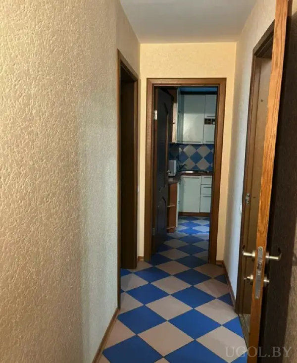 Узкий коридор с текстурными стенами и плиткой в сине-бежевую клетку, переходящей в кухонную зону с белыми шкафами и фартуком в том же стиле.