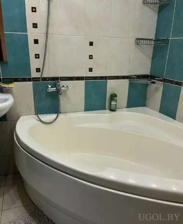 Угловая ванна с душем, стены отделаны бирюзовой и белой плиткой с декоративными вставками, металлические полки и коврик на полу.