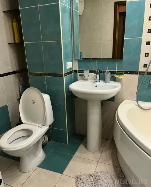 Светлая ванная с бирюзовой плиткой, раковиной с зеркалом, открытым унитазом и встроенными полками с бытовой химией.