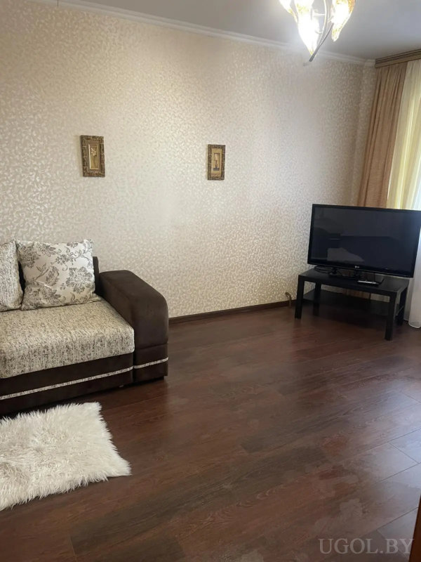 Светлая гостиная с диваном, белым ковром и телевизором на черной тумбе, оформленная в спокойных тонах.