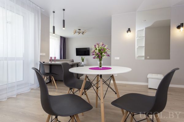 Комната с круглым столом, чёрными стульями, букетом на фиолетовой салфетке и серым диваном на фоне телевизора.