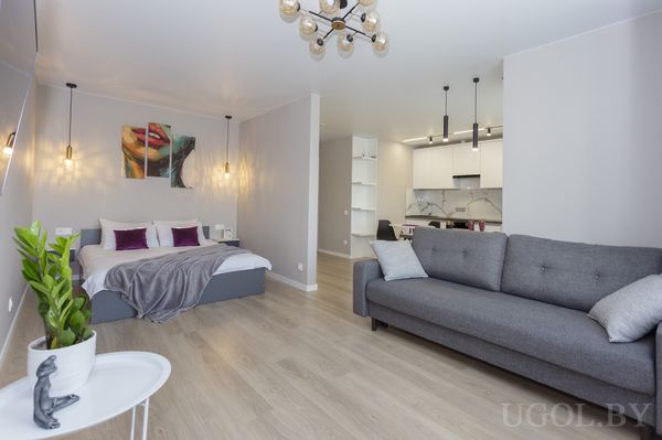 Открытая планировка с кроватью, диваном, белой тумбой, кухней с мраморным фартуком и подвесными светильниками.