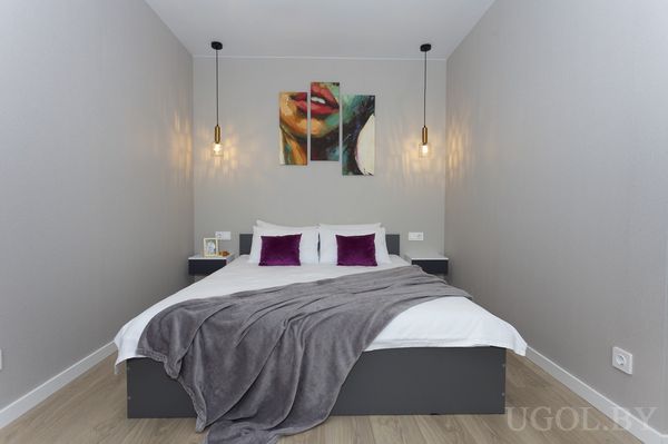 Комната с кроватью, серым покрывалом, фиолетовыми подушками и трёхпанельным панно с женским портретом.