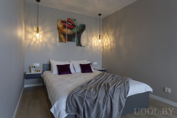 Интерьер с кроватью, фиолетовыми подушками, подвесными светильниками и панно с абстрактным лицом.