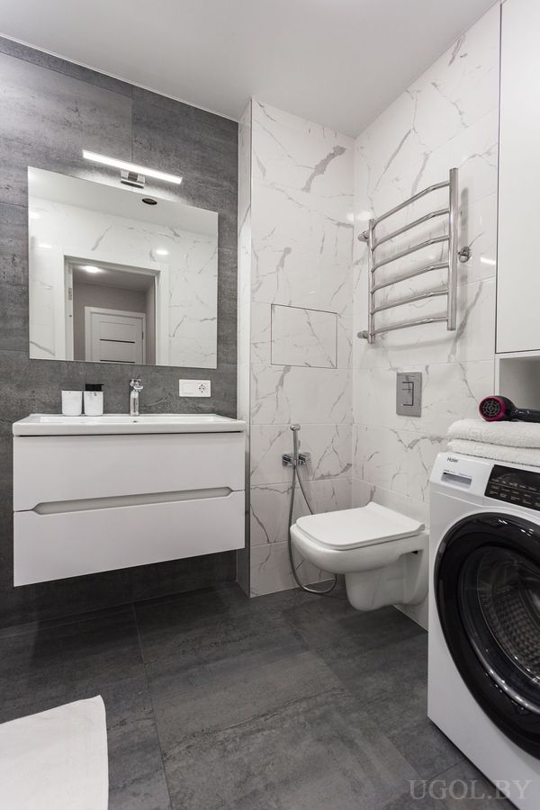Комната с белым умывальником, зеркалом с подсветкой, унитазом, машиной и плиткой с мраморным рисунком.