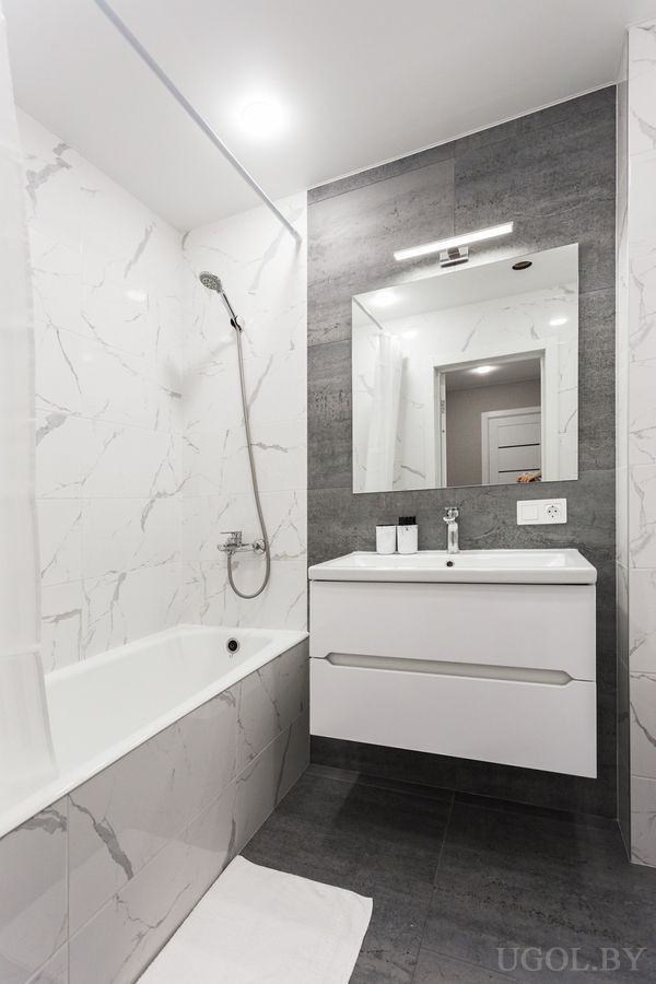 Интерьер с ванной, умывальником, зеркалом, тёмной плиткой и белым полотенцем на полу.